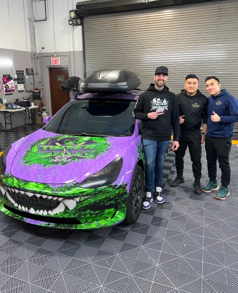Three men posing beside a Tesla Model 3 with a bold custom wrap featuring a green and purple Venom-style design, sharp teeth graphic on the front bumper, and "TUNER EVIL" lettering on the hood.
