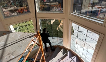 Worker applies window film to a large, multi-panel window inside a home with a curved staircase and high ceilings. Sunlight streams through the glass, casting reflections onto the floor and highlighting the view of a residential yard outside.