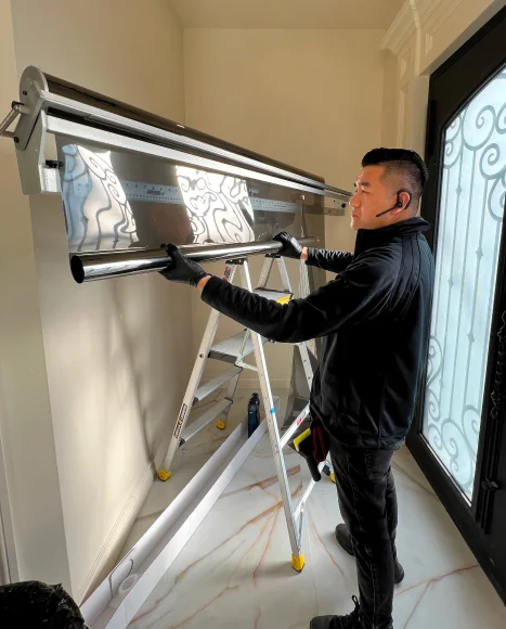 Man in black gloves and headset standing on a ladder, carefully aligning and cutting a roll of reflective window tint film using a large precision cutting machine indoors near a glass door.