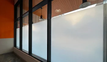 Large glass windows with frosted film applied to the lower two-thirds, providing privacy while still allowing natural light into the space. The interior features an orange and white wall and a wooden ceiling with hanging light fixtures.