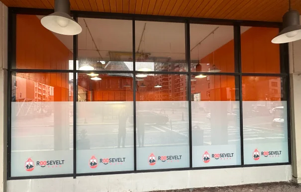 Storefront with large glass windows covered in frosted privacy film featuring repeated "Roosevelt Pups" logos along the bottom. The upper part of the windows reveals a brightly lit orange interior and a reflection of buildings across the street.