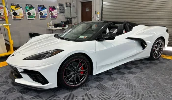 White Chevrolet Corvette convertible with a black interior parked inside a detailing garage with patterned flooring. The car features red brake calipers and black wheels, and several vinyl wrap color samples are displayed on the wall in the background.