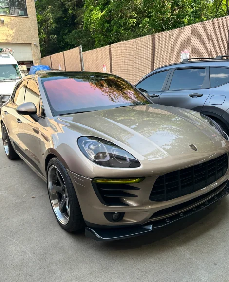 Customized tan Porsche SUV with a glossy finish, pink-tinted windshield, blacked-out accents, and aftermarket wheels parked in a lot beside a fence.