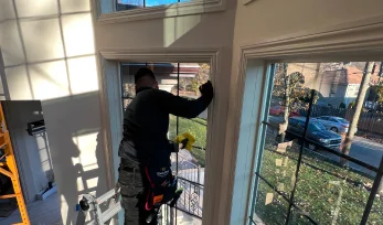 A technician stands on a ladder inside a bright home, applying window film to a tall window. Sunlight casts grid-like shadows on the wall, and a quiet suburban street with parked cars and trees is visible outside.