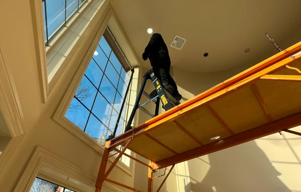 Worker standing on a ladder atop scaffolding while applying window film to a tall interior window in a high-ceilinged room. Bright sunlight streams through the glass, casting shadows on the beige walls.