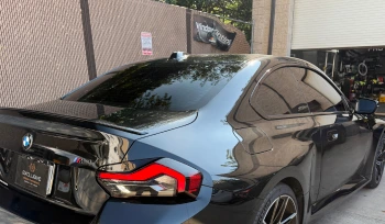 Rear and side view of a black BMW M Series coupe with dark tinted windows parked outside a window tinting shop. The reflection of trees is visible on the glossy surface of the car.