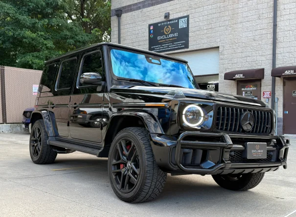 Glossy black Mercedes-Benz G-Class SUV with tinted windows, red brake calipers, and custom detailing, parked outside a shop with signage for Exclusive Window Tinting and autobahn window films.