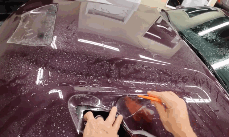 Close-up of a person applying paint protection film to the hood of a wet maroon car using a squeegee tool.