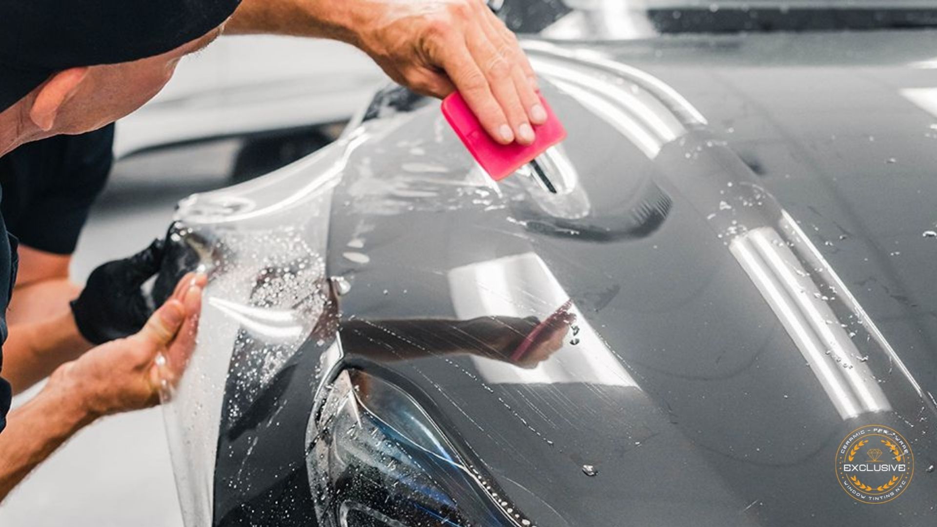 Technician smoothing paint protection film onto a gray car's hood with a red squeegee, surrounded by water droplets.