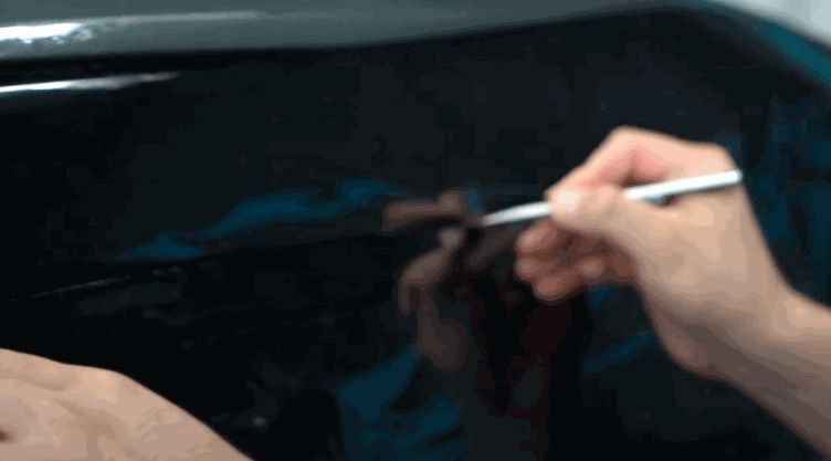 A person carefully trimming a piece of dark window tint film using a precision blade on a car window.