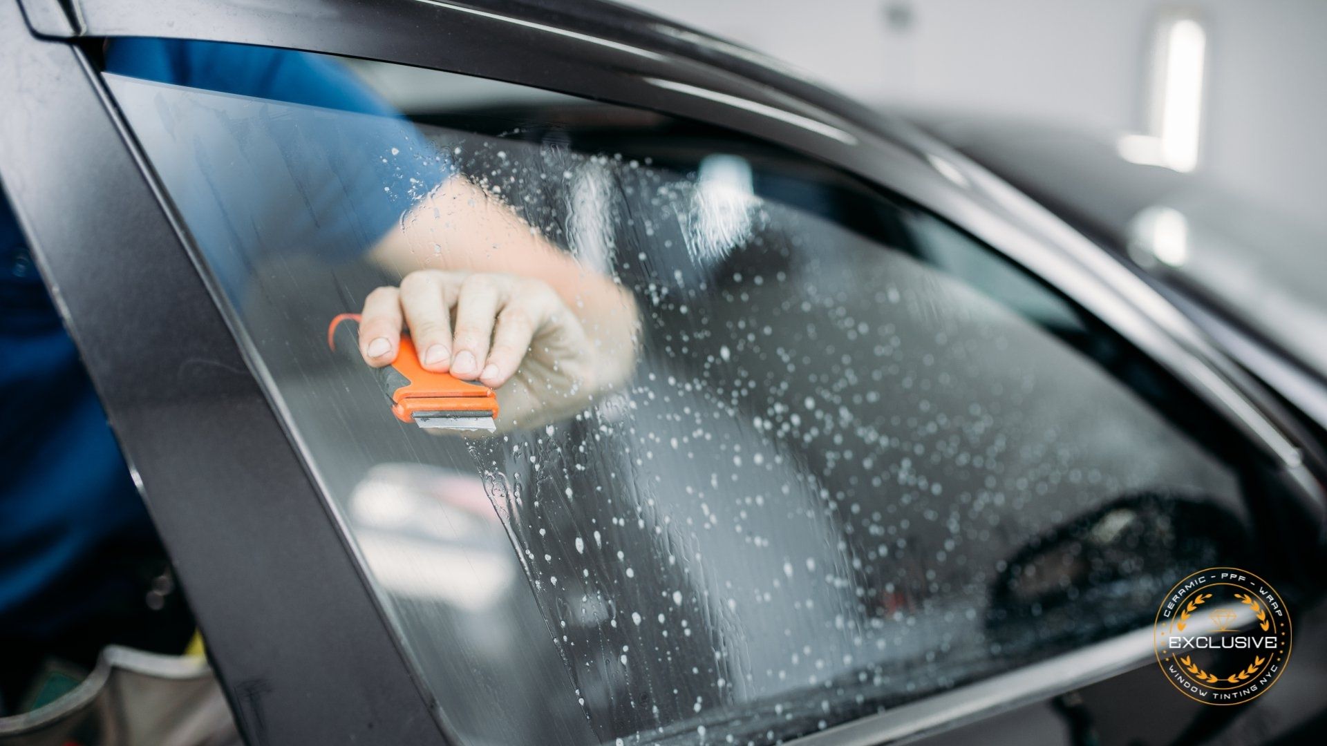 A close-up of a person using a scraper tool to clean a soapy car window during a window tinting preparation process.